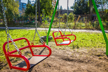 Colorful children playground activities in public park surrounded by green trees. Red children's swing