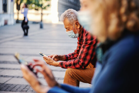 Senior an with protective mask on sitting on the bench outside and using cell phone. In foreground is senior woman using phone and wearing mask, too. Seniors esteem social distance.