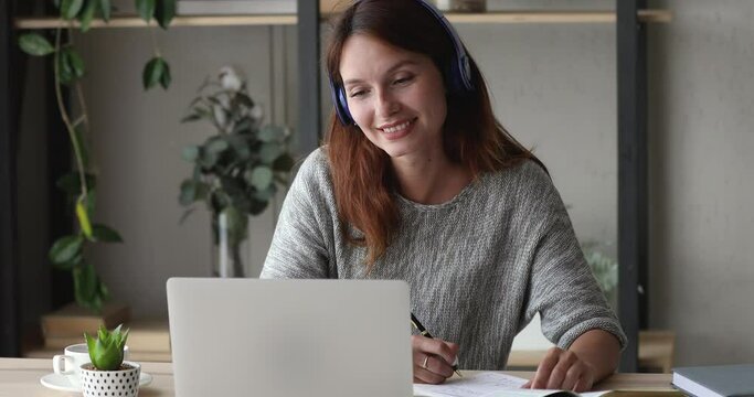 Smiling young caucasian smart woman student listening interesting online educational lecture, sitting at home workplace in wireless headphones, writing notes, distant e-learning education concept.