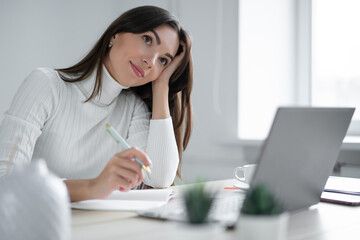 woman in glasses sits in front of laptop and holds pen in her hand.