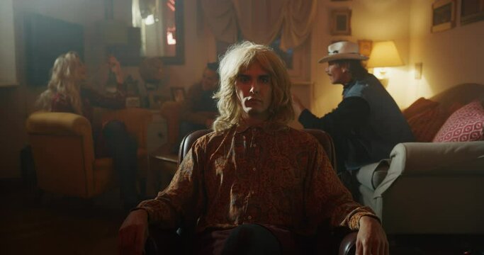 Cinematic shot of young carefree homosexual male gay is taking off wig and smiling in camera on a background of his friends are having fun. Concept of gender expression, identity and diversity.