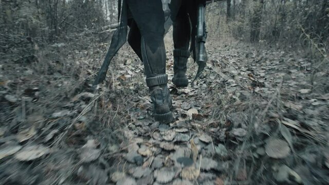 Close-up following shot of female legs step. Woman in futuristic military clothes feet walk along path in dead forest. Stalker concept, survivor with gun during war. Post apocalyptic world