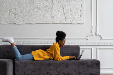 Smiling Afro-American biracial woman with afro hairstyle in yellow cardigan lying on couch, resting, looking at camera webcam and talking on video chat with her friends, watching webinar. - Powered by Adobe