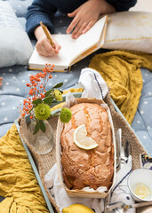 Closeup view of a delicious vegan lemon cake with a lemon slice on top. A blurred girl is writing in a diary at the back.