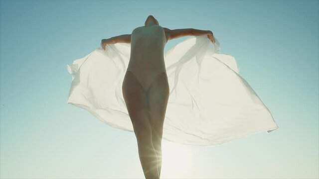 Flexible female gymnast or ballet dancer is dancing with huge silk fabric fluttering in wind on sky and sun flare background. Concept of tenderness, lightness, art and talent in nature