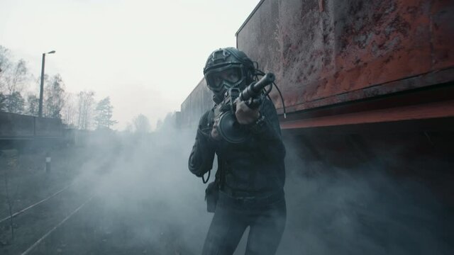 Female surviving future soldier aims with assault rifle and moves towards target by rusty train carriage. Lonely cyborg woman in futuristic combat suit goes through cloud of smoke during battle