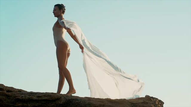 Flexible female gymnast or ballet dancer is dancing with huge silk fabric fluttering in wind on sky and sun flare background. Concept of tenderness, lightness, art and talent in nature