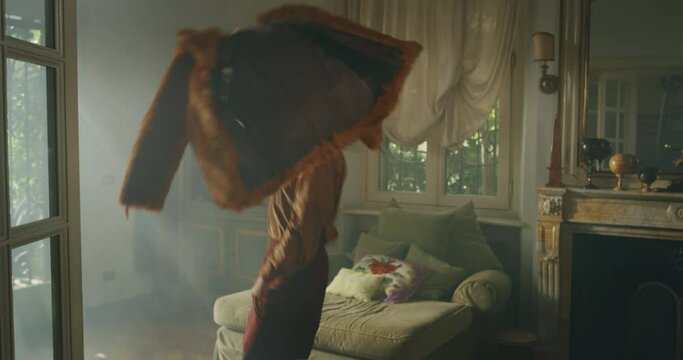 Cinematic shot of young carefree happy smiling transgender woman wearing sunglasses, wig and fur coat is having fun to listening to the music, dancing crazy and jumping on sofa in living room at home.