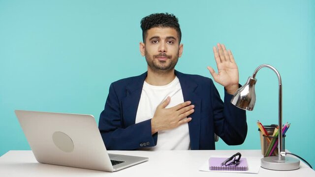 Serious businessman in suit swearing to say truth, sitting on his workplace raising one hand and holding on chest another, promise. Indoor studio shot isolated on blue background