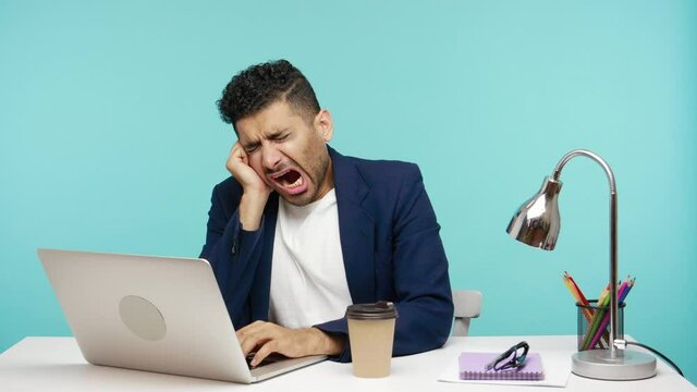 Tired extremely exhausted businessman yawning working on laptop at his office until late, checking paper coffee cup, overwork. Indoor studio shot isolated on blue background