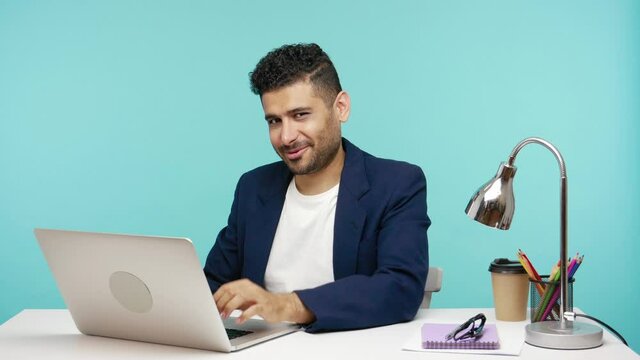 Handsome playful businessman saying come here calling with finger and looking at camera with smile, working on laptop in office, flirt, workplace affair. Indoor studio shot isolated on blue background