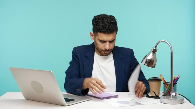 Serious responsible businessman looking for proper documents and data preparing report at laptop sitting in his office. Indoor studio shot isolated on blue background