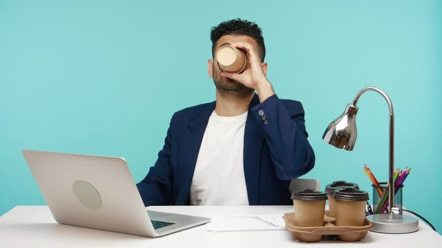 Tired busy employee working on laptop drinking lot of coffee, lack of energy and inspiration in morning, deadline at work. Indoor studio shot isolated on blue background