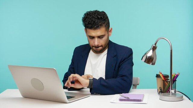 Busy office worker typing on laptop sitting at his workplace, checking time and daily goals completion on smartwatch on his wrist. Indoor studio shot isolated on blue background