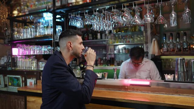 Confidence Caucasian man sitting at bar counter enjoy drinking cocktail and talking with barman in nightclub. Asian male mixologist bartender preparing mixed alcohol drink on rocks glass for customer