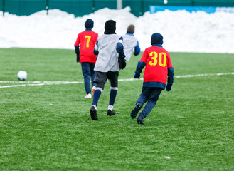 Boys in red sportswear running on soccer field with snow on background. Young footballers dribble and kick football ball in game. Training, active lifestyle, sport, children winter activity