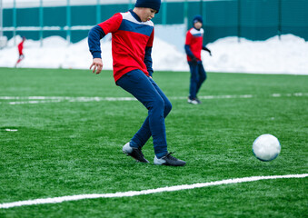 Boys in red sportswear running on soccer field with snow on background. Young footballers dribble and kick football ball in game. Training, active lifestyle, sport, children winter activity