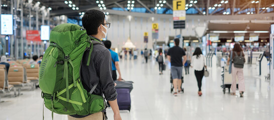 Young man traveler wearing medical face mask and checking flight time in airport terminal,...