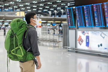 Young man traveler wearing medical face mask and checking flight time in airport terminal,...