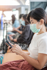 Obraz na płótnie Canvas Young female wearing face mask and using mobile smartphone in airport, protection Coronavirus disease (Covid-19) infection, Asian woman traveler sitting on chair. New Normal and social distancing