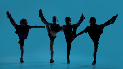 Silhouetted full length shot of four little kids, gymnasts showing flexibility while posing, standing isolated over blue background