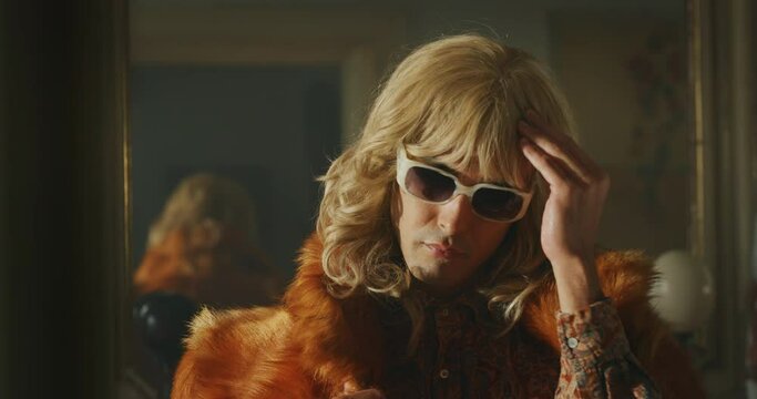 Cinematic shot of young carefree happy smiling transgender woman wearing wig and fur coat is taking off sunglasses and smiling in camera at home. Concept of gender expression, identity and diversity.