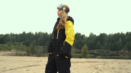 A girl in overalls and glasses in the summer looks at the lake holding a snowboard in her hands