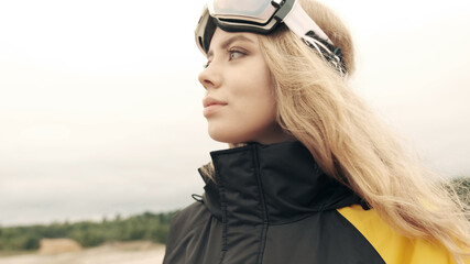 A girl in overalls and glasses in the summer looks at the lake holding a snowboard in her hands