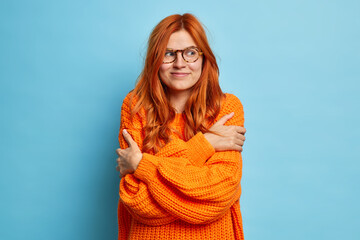 Nice looking redhead woman embraces herself likes her new knitted orange sweater looks satisfied on right poses against blue background. Lovely pretty millennial girl dressed in comfortable jumper