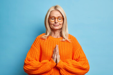 Peaceful blonde woman keeps eyes closed stands in praying pose prays for good wellness dressed in...