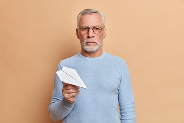 Serious senior man with thick beard holds paper air plane looks confidently at camera holds paper airplane wears optical glasses casual jumper poses over beige wall. Self assured mature businessman