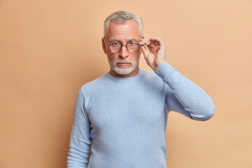Self confident serious man with grey beard keeps hands on glasses looks directly at camera dressed in casual jumper listents information carefully poses against beige background. Hadsome retired male