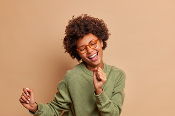 Fototapeta na wymiar Feeling boost of happiness. Carefree Afro American woman sings favorite song and dances joyfully to rhythm closes eyes with pleasure smiles broadly has white teeth isolated over brown background