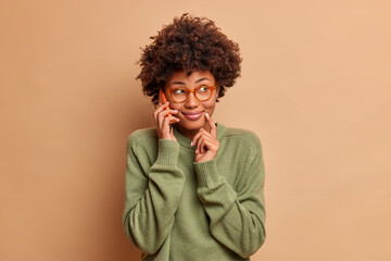 Fototapeta na wymiar Image of pretty smiling woman talks on mobile phone with friend discuss plans for weekends concentrated aside happily has smart look wears transparent eyewear casual sweater. Technology lifestyle