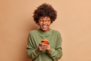 Fototapeta na wymiar Portrait of good looking woman uses mobile phone receives pleasant message smiles broadly and looks thoughtfully aside reads feedback under her post in social networks isolated on beige wall