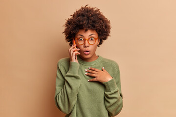 Fototapeta na wymiar Astonished curly haired woman talks on phone learns terrible event happened holds smartphone near ear stands with bated breath wears transparent glasses and sweater isolated over beige background