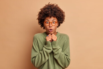 Fototapeta na wymiar Emotional shocked Afro American woman with curly hair holds breath keeps hands together opens mouth with amazement stares bugged eyes dressed in casual jumper and optical spectacles models indoor