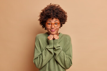 Fototapeta na wymiar Pleased cheerful female student with Afro hair keeps hands under chin dressed in casual jumper wears glasses has charming smile on face isolated over beige background. Face expressions concept