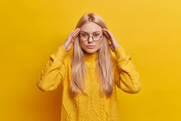 Cute serious light haired young woman keeps hands on temples tries to concentrate has headache...