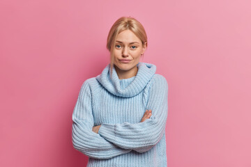 Serious self confident young blonde adorable European woman listens interlocutor with dissatisfied expression accepts explanations from husband wears blue sweater models against pink background.