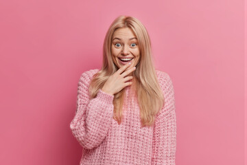 Surprised amused blonde woman stares at something wonderful with broad smile looses speech from amazement dressed in warm winter sweater isolated over rosy background cannot believe in her achievement