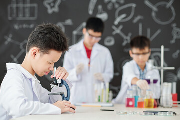 Curious Asian schoolboy looking though microscope at desk in science class