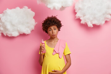Upset dark skinned pregnant woman holds tummy suffers contractions being on third trimester holds feeding bottle carries baby bodysuit on shoulder poses indoor. Pregnancy and expectation concept