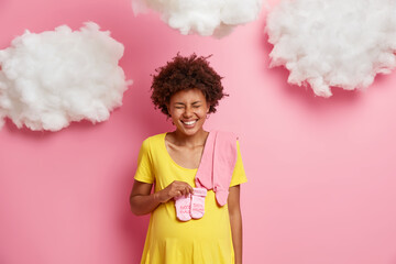 Overjoyed dark skinned woman with Afro hair holds small babys socks over belly laughs happily and keeps eyes closed anticipates for newborn isolated over pink background with white fluffy clouds
