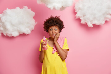 Photo of worried nervous pregnant ethnic woman bites finger nails and looks anxiously at camera holds milk bottle poses with baby stuff against rosy background has big stomach. Maternity concept