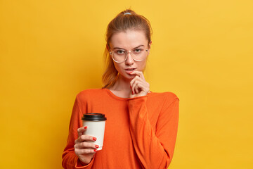 Charming serious Caucasian girl looks seriously at camera drinks coffee to go wears orange jumper and big round spectacles isolated over yellow background. Teenager has break between lessons