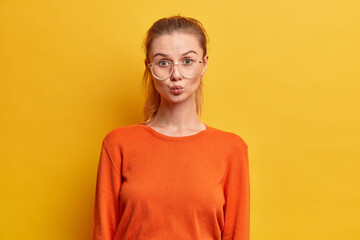 Horizontal shot of good looking woman with folded lips wants to kiss someone has flirty expression wears big transparent glasses orange jumper isolated over yellow background. Romantic feelings