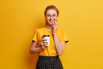 Isolated shot of cheerful girl smiles happily expresses sincere feelings drinks takeaway coffee hears something positive from interlocutor wears round eyewear isolated over vivid yellow background