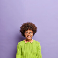 Vertical shot of happy curly haired young woman smiles broadly concentrated above sees something incredible looks upwards wears round spectacles and green jumper poses against purple background