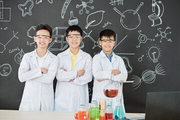 Group of happy schoolchildren in labcoats and protective goggles folding arms and smiling at camera